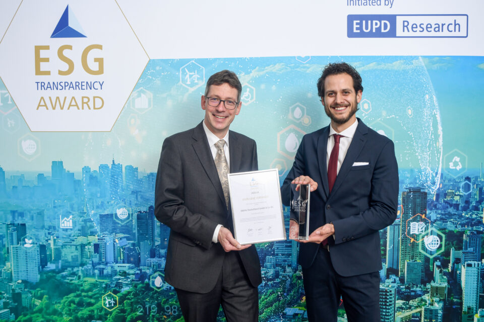 Zwei Männer in Anzügen stehen vor einer ESG Transparency Award-Wand, einer hält eine Urkunde, der andere eine Trophäe