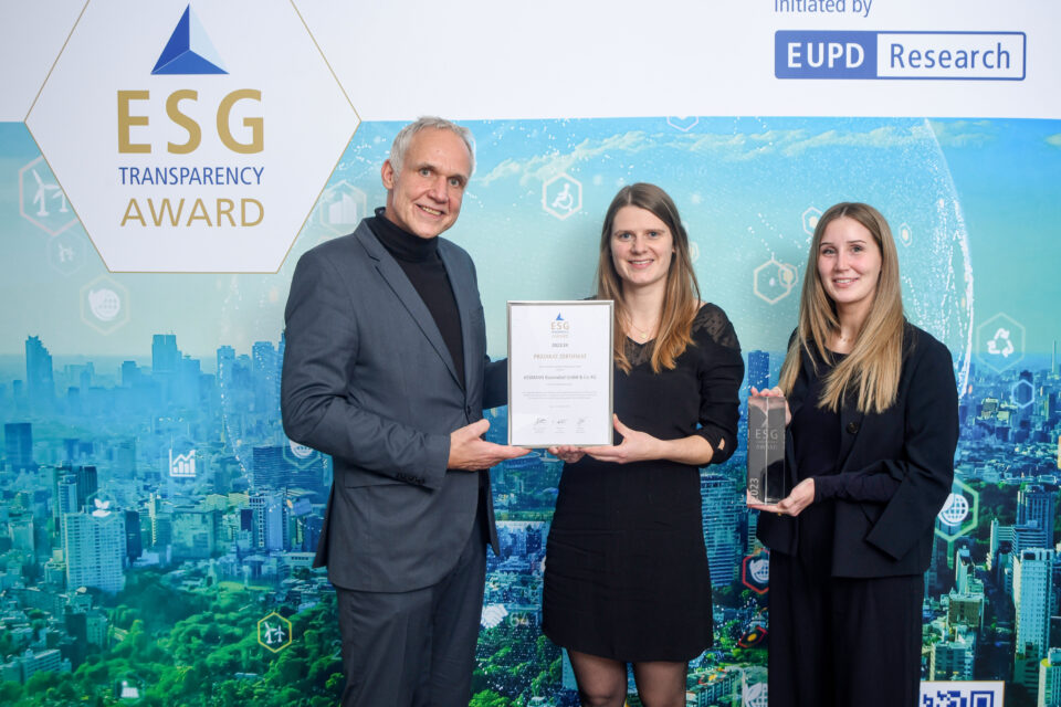 Drei Personen stehen vor einer ESG Transparency Award-Wand, zwei Frauen und ein Mann, die Frau in der Mitte hält eine Urkunde, die Frau rechts hält eine Trophäe.