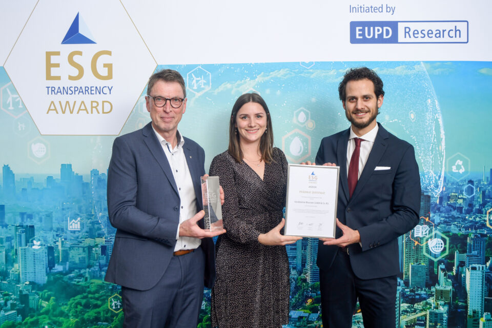 Drei Personen in formeller Kleidung stehen vor einer Wand mit dem Logo des ESG Transparency Award und halten eine Trophäe sowie eine Urkunde