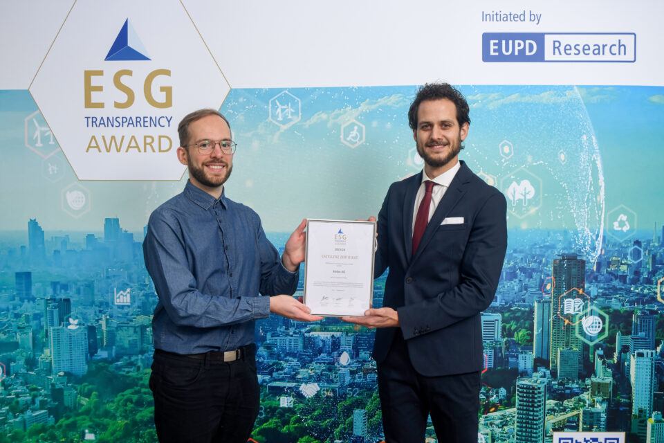 Zwei Männer stehen vor einer großen digitalen Stadtlandschaft, halten gemeinsam eine Urkunde mit dem ESG Transparency Award-Logo