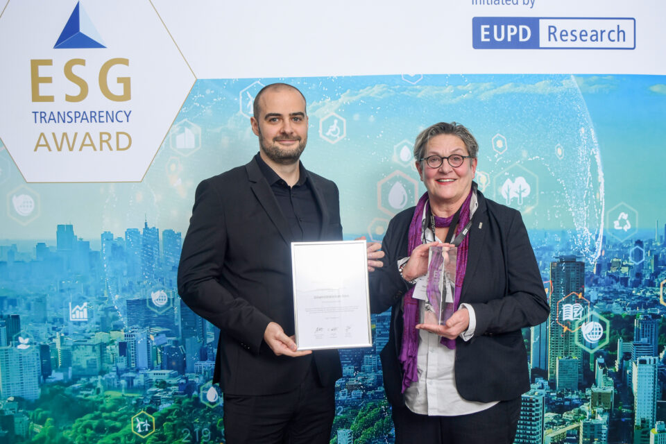 Zwei Personen stehen vor einer Wand mit dem Schriftzug 'ESG Transparency Award' und halten eine Urkunde sowie eine Trophäe